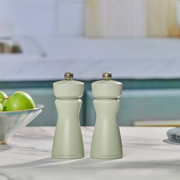 Two Cole & Mason Kenton Sage Salt & Pepper Mills with a matte light green finish stand side by side on the kitchen counter, featuring a ceramic mechanism. Nearby is a bowl of green apples against a softly blurred blue and white background.