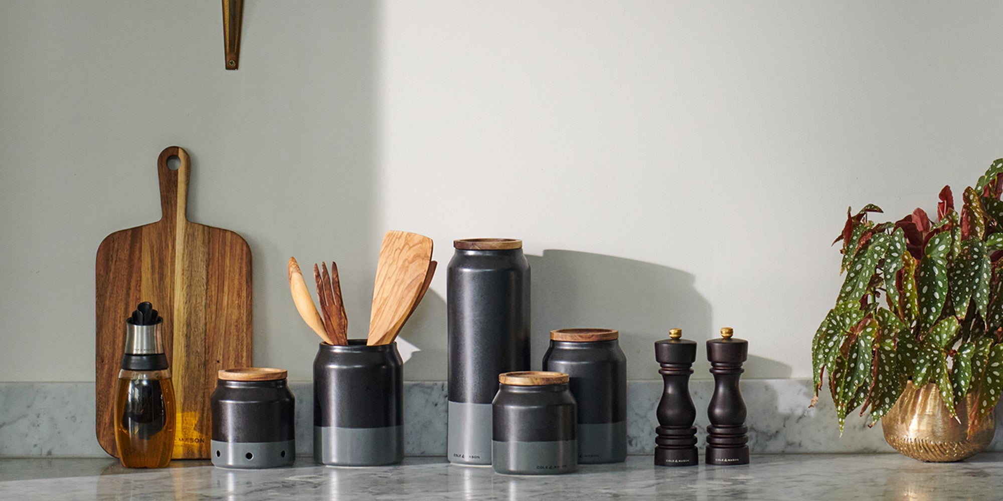 A kitchen countertop with black and gray canisters, wooden utensils, two pepper mills, a potted plant, an oil dispenser, and a wooden cutting board against a white wall.