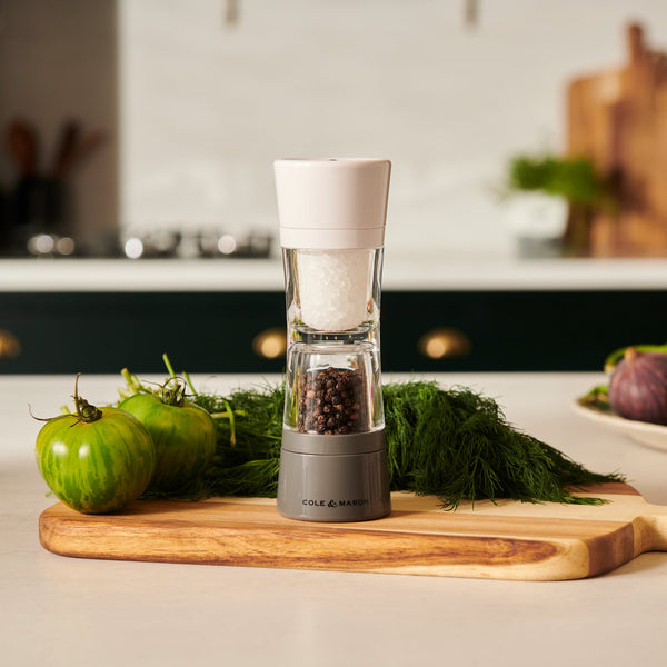 The Cole & Mason Lincoln 2-in-1 Duo Mill with adjustable settings rests on a wooden cutting board beside fresh green tomatoes and dill, set in a modern kitchen with blurred background elements.