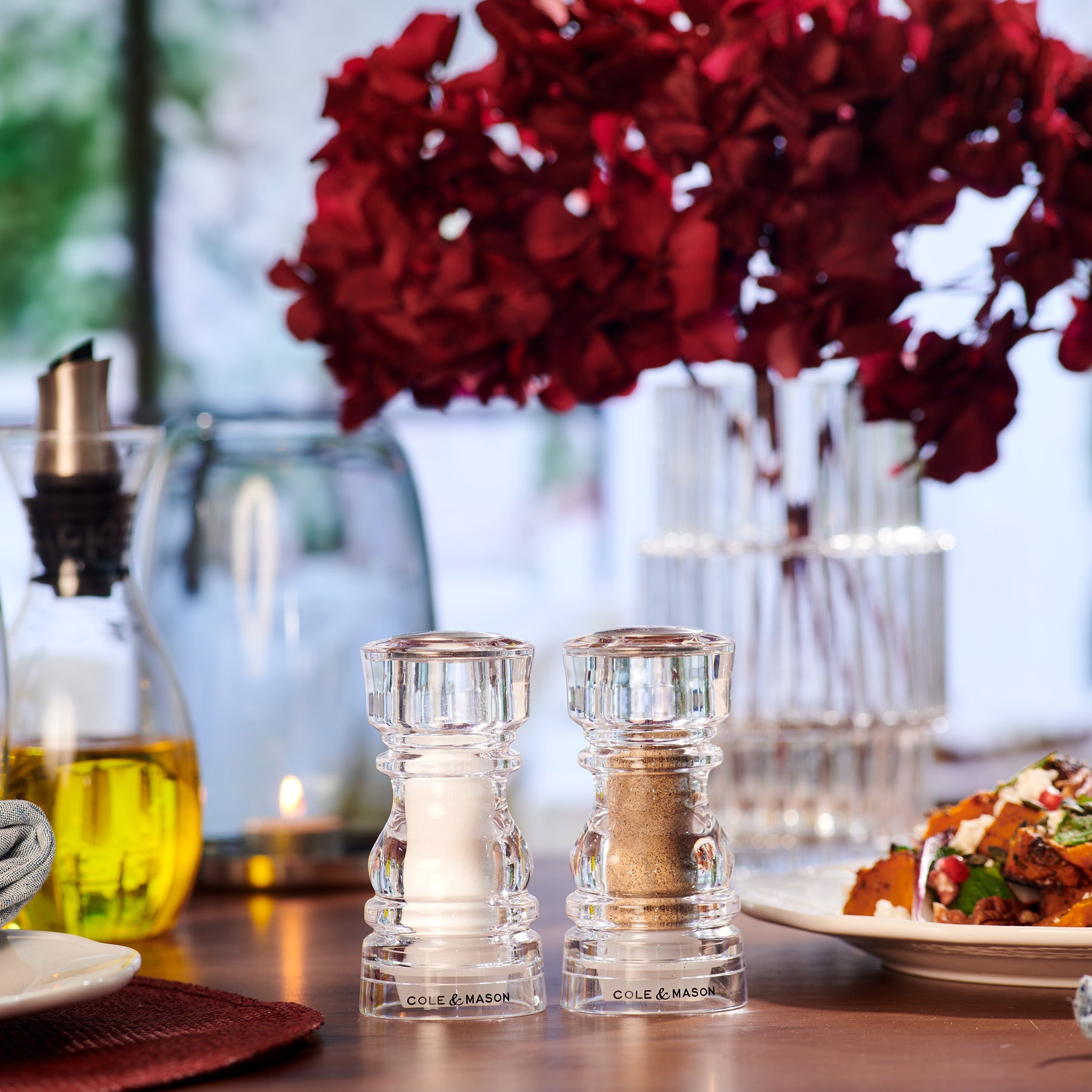 #130mm A pair of Cole & Mason London Acrylic Salt & Pepper Shakers are displayed on a dining table beside a vase of red flowers, an oil bottle, and a plate of food in an elegantly lit setting.