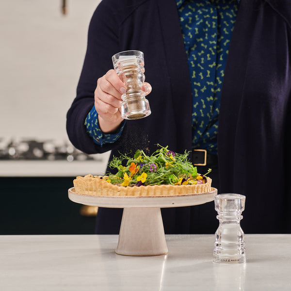 #130mm A person sprinkles pepper from a Cole & Mason grinder onto a tart with fresh greens and edible flowers on a pedestal, while the Cole & Mason London Acrylic Salt & Pepper Shaker sits on the counter nearby.
