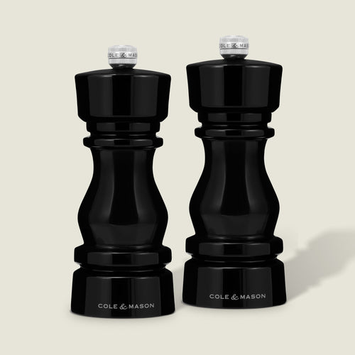 Cole & Mason’s London Black Acrylic Salt & Pepper Mills (150mm) stand side by side on a beige background, featuring glossy black curves, metallic tops, and precision grinding with adjustable settings.