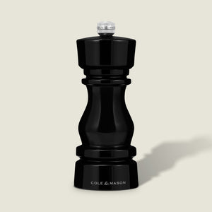 The Cole & Mason London Black Acrylic Salt & Pepper Mills 150mm features a glossy hourglass design and premium grinding technology, shown standing upright against a light beige background.
