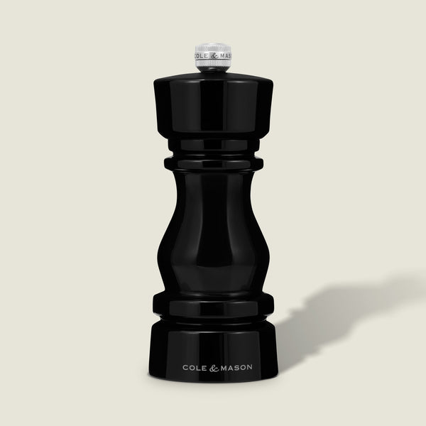 The Cole & Mason London Black Acrylic Salt & Pepper Mills 150mm features a glossy hourglass design and premium grinding technology, shown standing upright against a light beige background.
