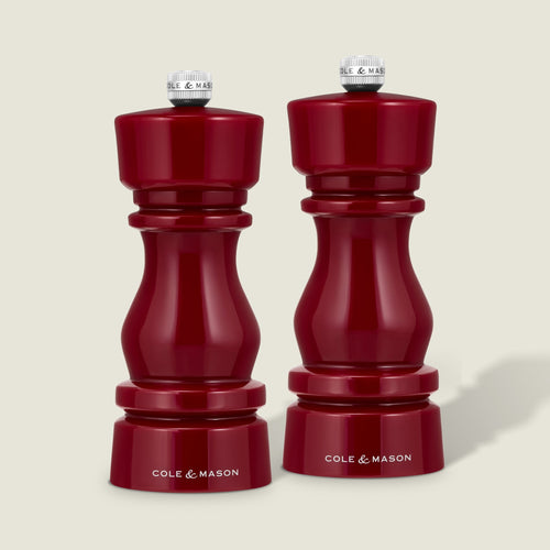 A pair of Cole & Mason London Red Acrylic Salt & Pepper Mills (150mm) stand side by side on a light background, each featuring a glossy red finish, elegant modern curves, silver top knobs, and adjustable grinding mechanisms for customized seasoning.