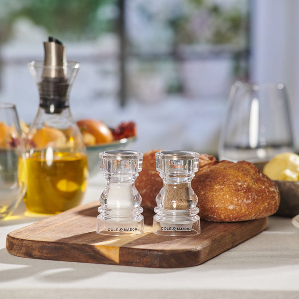 #86mm 
A wooden board holds Cole & Mason London Acrylic Salt & Pepper Shakers. Behind them are bread rolls, butter, an oil dispenser, and glasses on a set dining table with a blurred outdoor background.