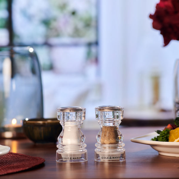 #86mm
Two clear Cole & Mason London Acrylic Salt & Pepper Shakers sit on a wooden dining table set for a meal, with plates of food and a candle in the background.