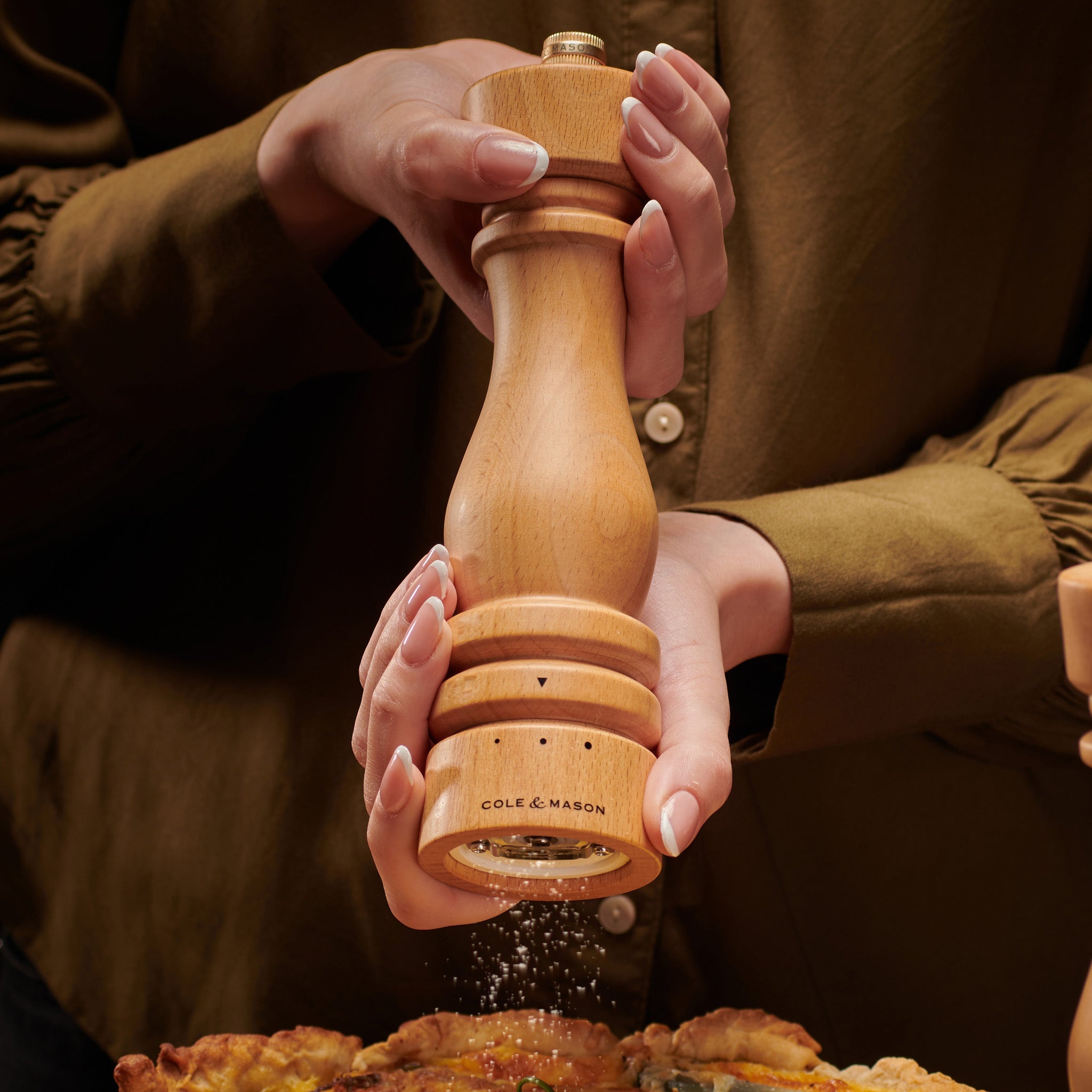 A person wearing a brown shirt holds a wooden Cole & Mason pepper grinder over food, sprinkling pepper onto a dish.