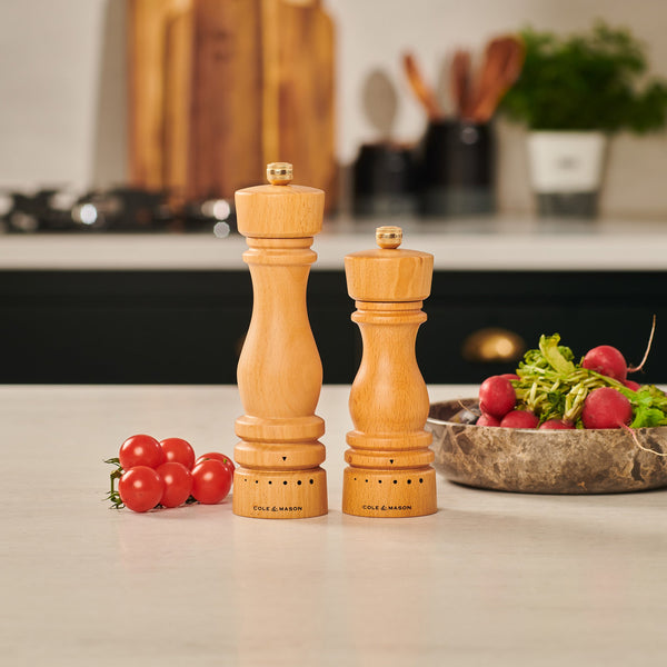 Two Cole & Mason UK London Natural Beech Grind Select Salt & Pepper Mills stand on a kitchen counter near cherry tomatoes and a bowl of radishes and greens, with a softly blurred kitchen backdrop highlighting their elegant design.