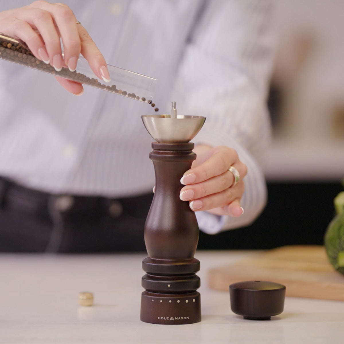 A person pours whole peppercorns into a dark wooden pepper grinder in a kitchen, with a cutting board and vegetables blurred in the background.