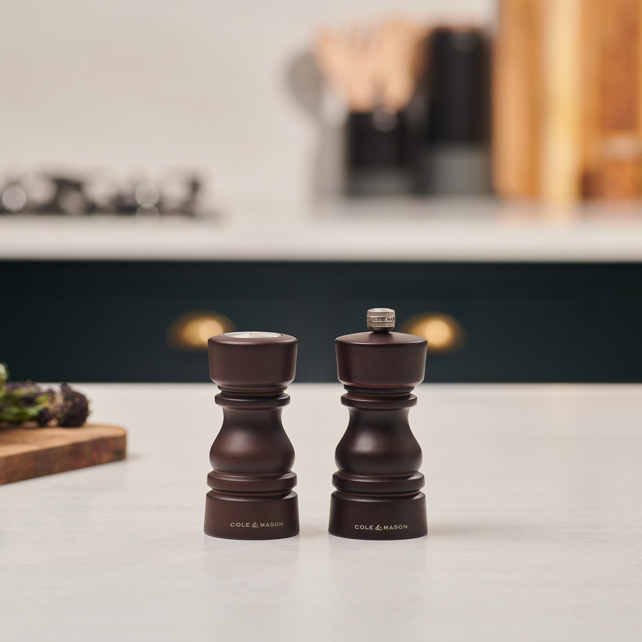 The Cole & Mason London Chocolate Wood Salt Shaker & Pepper Mill Set sits side by side on a white kitchen countertop, crafted from natural beech wood with a rich dark finish. Blurred utensils and kitchen items are visible in the background.