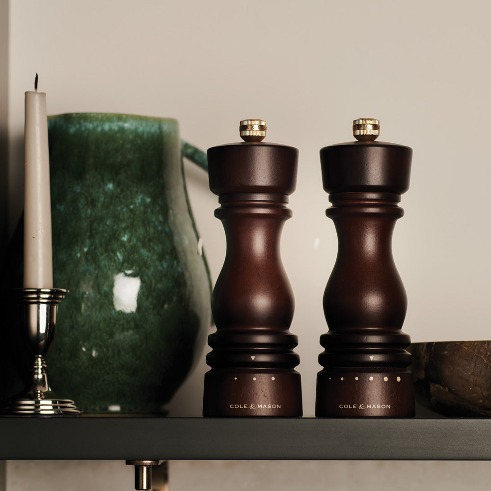 The Cole & Mason London Stained Wood Grind Select Salt & Pepper Mills, with a rich beech wood finish, sit side by side on a shelf near a green ceramic pitcher and white candle, ready to add customizable seasoning at your table.
