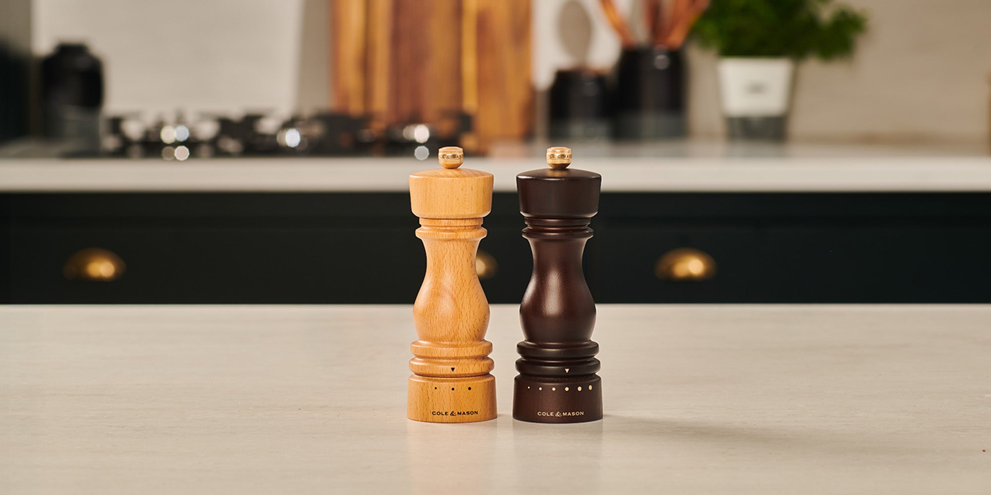 Two wooden mills, one light and one dark, are placed side by side on a kitchen counter with a modern kitchen background. The light one is likely for salt and the dark one is likely for pepper.