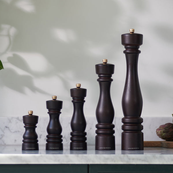 Five Cole & Mason London Chocolate Wood Salt & Pepper Mills of varying heights stand in ascending order on a marble countertop, backed by a white wall with soft natural light and plant shadows. Premium grinding technology ensures effortless use.