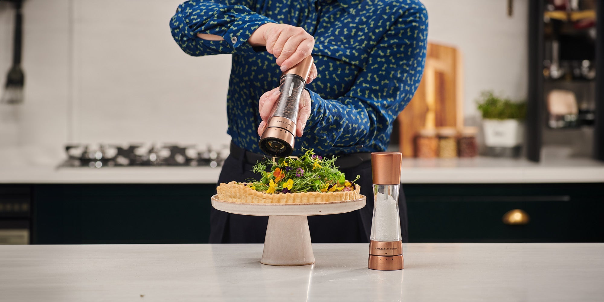 A person in a blue patterned shirt grinds pepper onto a leafy green salad tart placed on a cake stand in a modern kitchen, with a salt grinder on the counter.