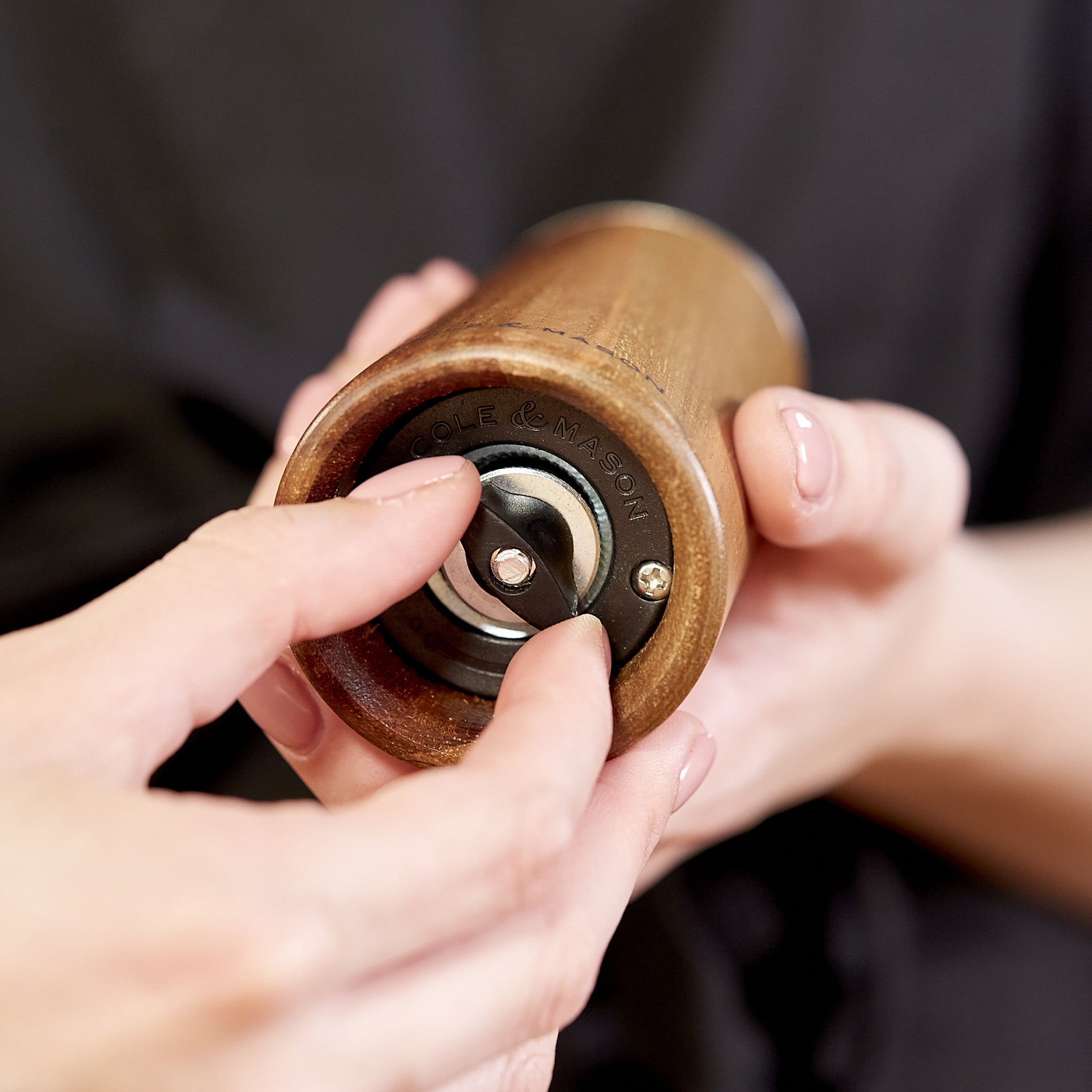 A person adjusts the grind setting on the bottom of a wooden pepper mill, holding the mill in one hand and turning the metal knob with the other.