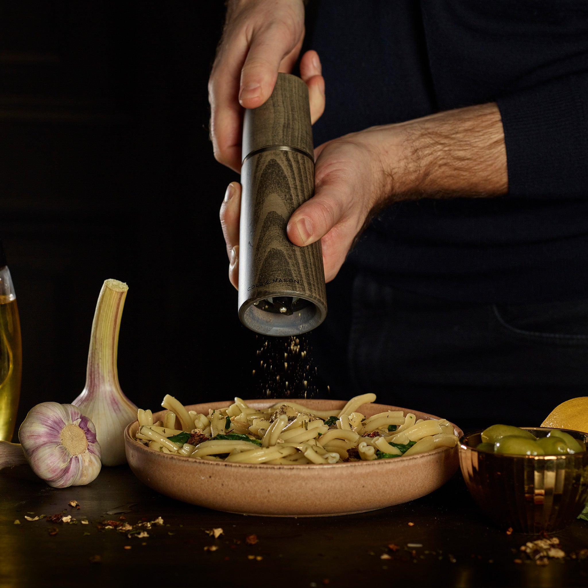 A person grinds black pepper onto pasta using Cole & Mason UK Marlow Grey Beech Salt & Pepper Mills with an adjustable grinder. Garlic, oil, and olives on the table stand out against a dark background for a modern look.