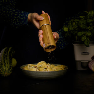 Using the Cole & Mason UK Marlow Natural Acacia Salt & Pepper Mill, someone sprinkles fresh pepper onto pasta shells, surrounded by vegetables and a basil plant on a dark table.