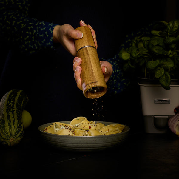 Using the Cole & Mason UK Marlow Natural Acacia Salt & Pepper Mill, someone sprinkles fresh pepper onto pasta shells, surrounded by vegetables and a basil plant on a dark table.