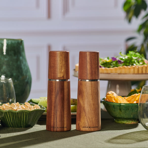 Two Marlow Natural Acacia Salt & Pepper Mills by Cole & Mason UK, featuring adjustable grinding mechanisms, sit on a green table with bowls of food and glasses, delivering precise seasoning in a stylish dining setting.