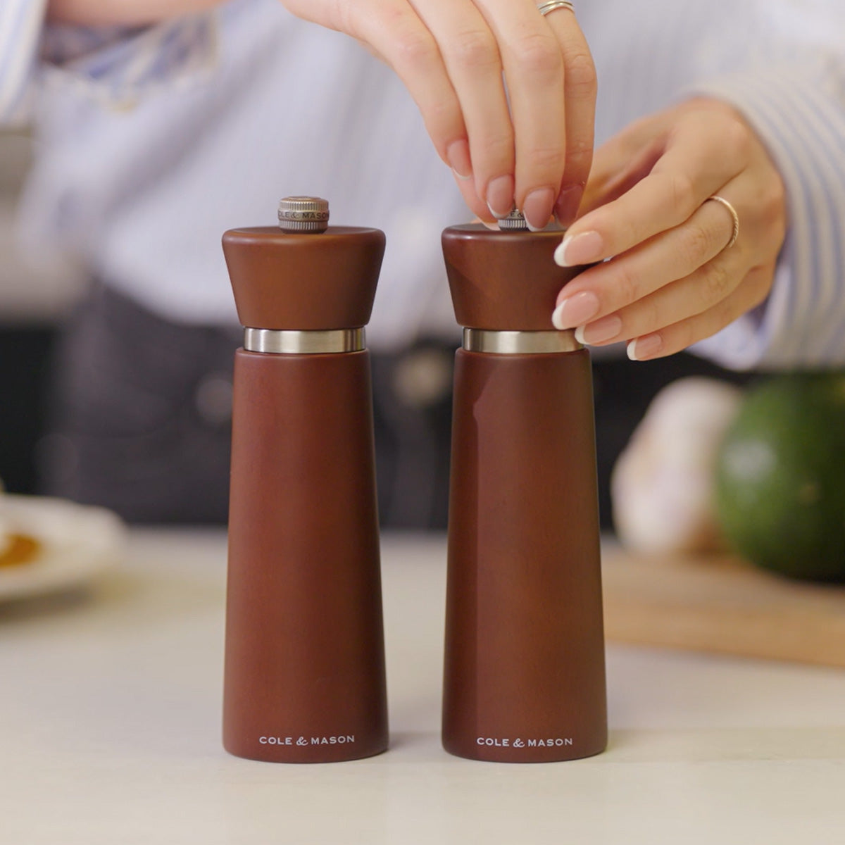 Two hands are adjusting and holding brown wooden salt and pepper grinders labeled Cole & Mason on a kitchen counter, with blurred food and produce in the background.