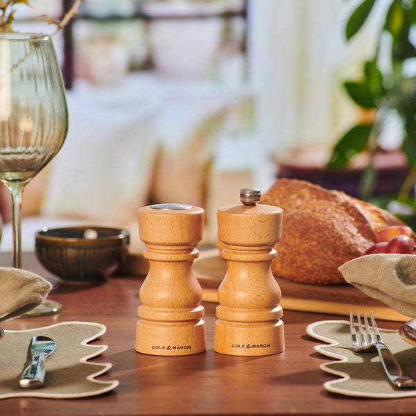 The Cole & Mason London Natural Beech Salt Shaker & Pepper Mill Set is displayed on a dining table set with glassware, utensils, napkins, bread, and greenery in the background.