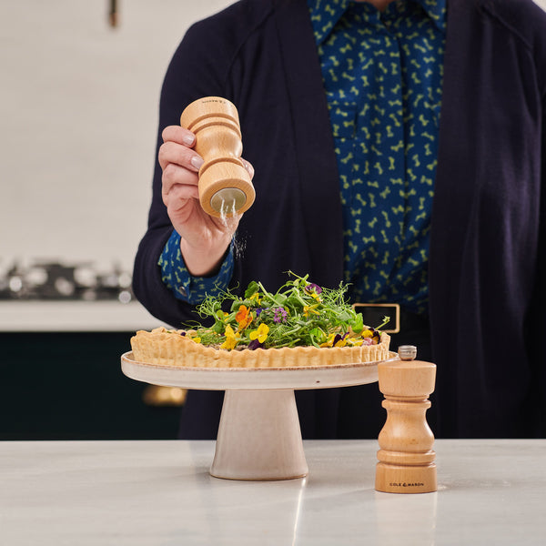 Someone seasons a savory tart with the Cole & Mason London Natural Beech Salt Shaker & Pepper Mill Set, garnished with fresh greens and edible flowers, all presented on a pedestal stand in a modern kitchen.