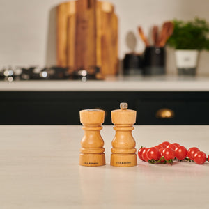 The Cole & Mason London Natural Beech Salt Shaker & Pepper Mill Set features Grind Select settings and sits on a kitchen countertop near cherry tomatoes, with cutting boards, utensils, and a potted plant blurred in the background.