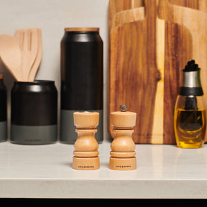 The Cole & Mason London Natural Beech Salt Shaker & Pepper Mill Set sits on a kitchen counter, surrounded by black containers, wooden utensils, a bottle of oil, and cutting boards in the background.