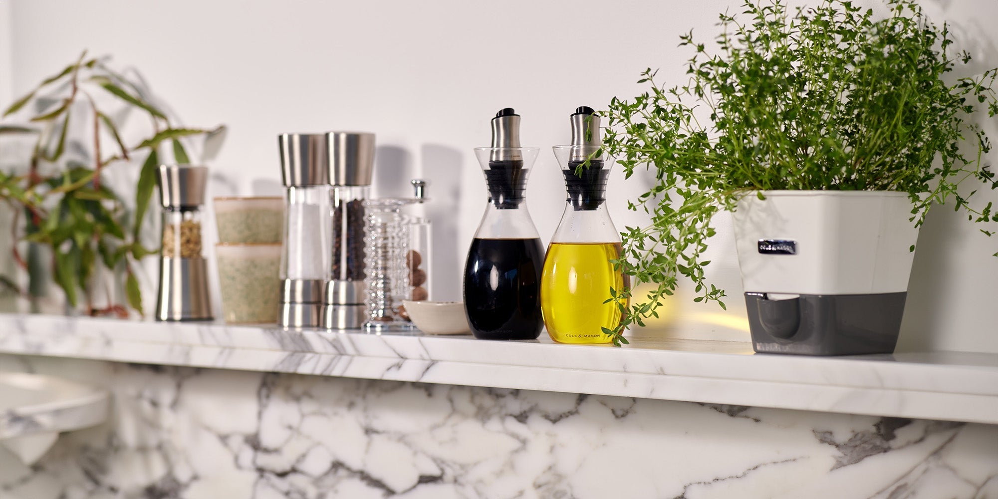 A marble kitchen shelf with spice grinders, glass oil and vinegar dispensers, a small bowl, and a potted green herb plant arranged neatly against a white wall.
