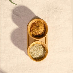 The Cole & Mason Portobello Olive Wood Pinch Pot Set features two small bowls, one filled with a brown spice blend and the other with white grains, their fine olive wood patterns shown on a textured surface.