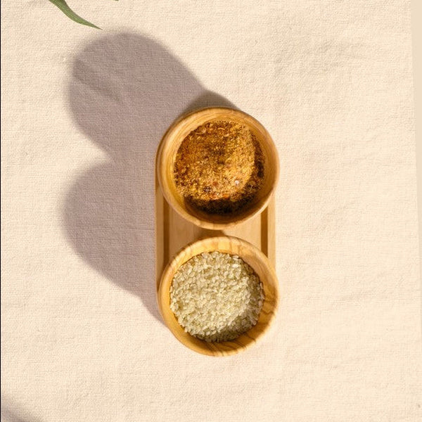 The Cole & Mason Portobello Olive Wood Pinch Pot Set features two small bowls, one filled with a brown spice blend and the other with white grains, their fine olive wood patterns shown on a textured surface.