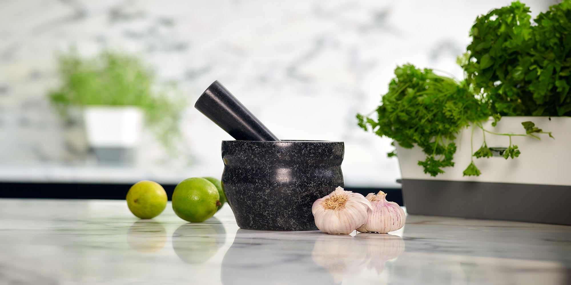 A stone mortar and pestle sits on a marble countertop, surrounded by two whole limes, two garlic bulbs, and a container of fresh green herbs. The background is softly blurred.