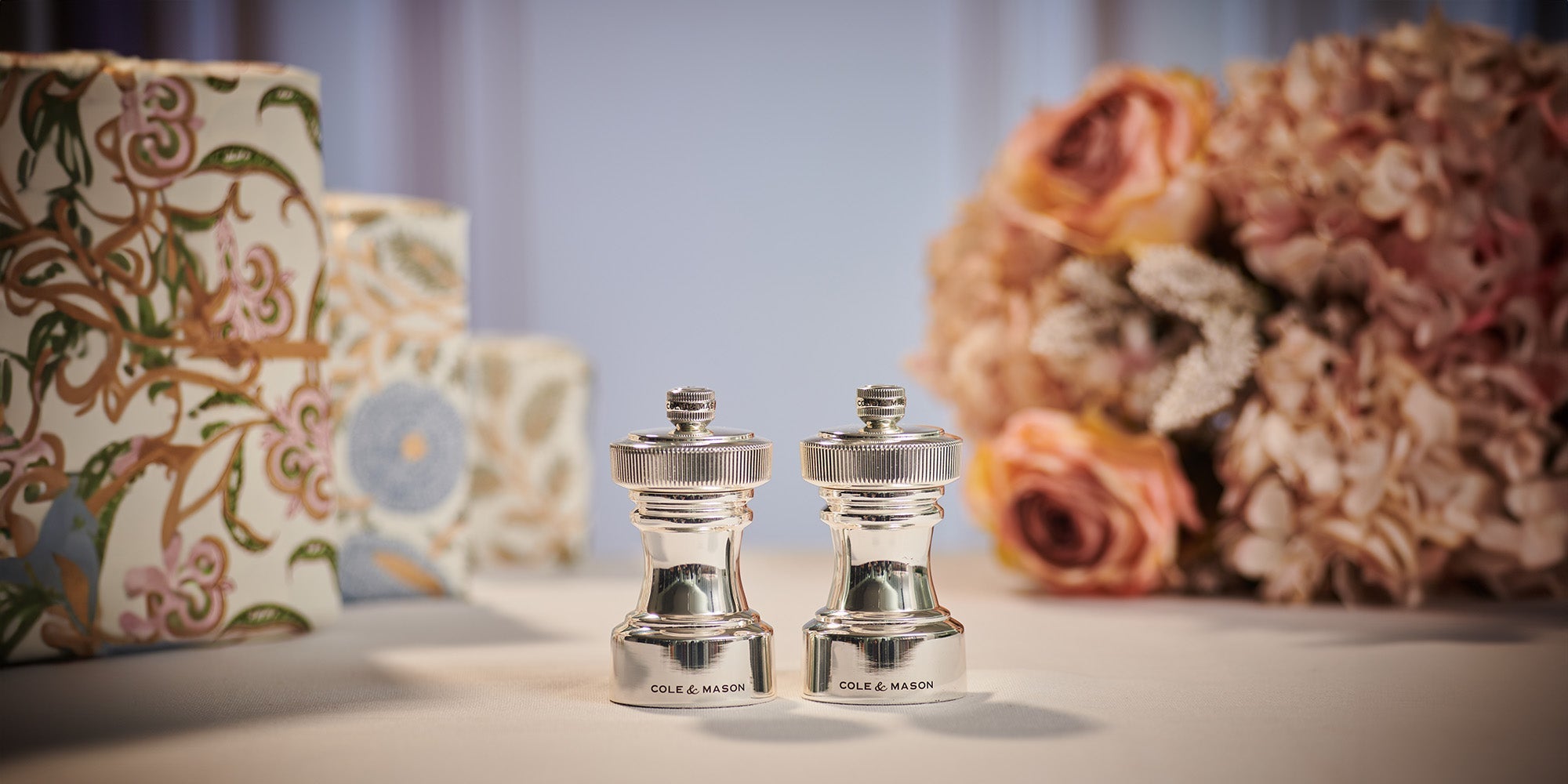 Two silver salt and pepper grinders labeled Cole & Mason are placed on a tabletop, with floral-patterned boxes and a bouquet of light pink and peach flowers in the softly blurred background.