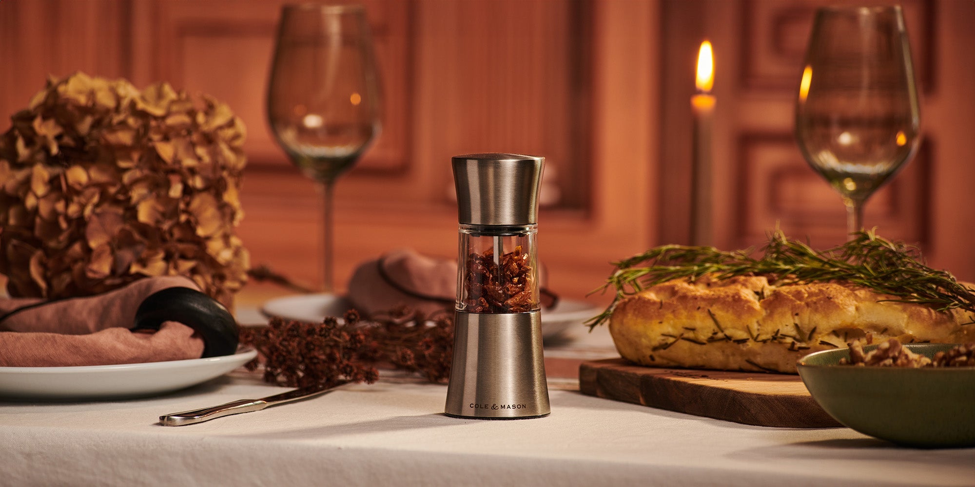 A table set for dinner features a pepper grinder, a loaf of bread with herbs, wine glasses, plates, cutlery, a bowl of walnuts, a candle, and dried flowers, all on a white tablecloth in warm lighting.