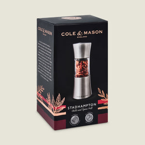 A black and red box showcases the Cole & Mason Stadhampton Chilli & Spice Mill with an ergonomic design, visible red spices in a clear section, precision grinding mechanism details, and brand logos on the packaging.
