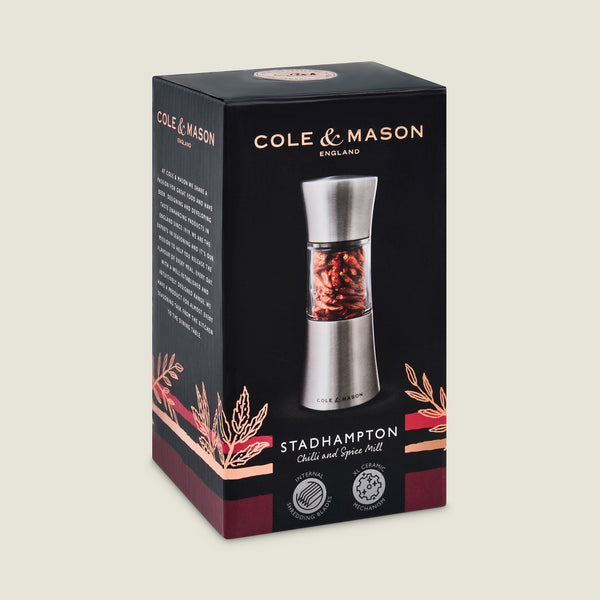 A black and red box showcases the Cole & Mason Stadhampton Chilli & Spice Mill with an ergonomic design, visible red spices in a clear section, precision grinding mechanism details, and brand logos on the packaging.