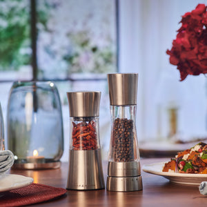 Two Cole & Mason Stadhampton Chilli & Spice Mills, featuring an ergonomic design and premium grinding mechanism, one with dried chili and one with black peppercorns, rest on a wooden table in a softly lit room set for dining.