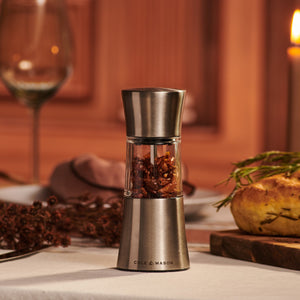 The Cole & Mason Stadhampton Chilli & Spice Mill, featuring an ergonomic design and stainless steel grinder, sits on a set table with red pepper flakes; a baked potato, fresh herbs, wine glass, and lit candle enhance the inviting scene.