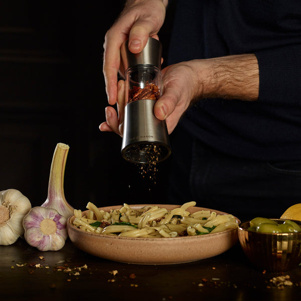 A Cole & Mason Stadhampton Chilli & Spice Mill grinds red pepper flakes over pasta, while fresh garlic, an olive-filled bowl, and a lemon wedge rest on a dark table nearby.