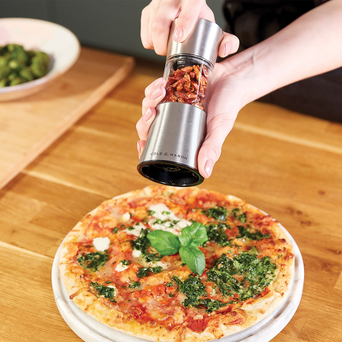 A hand holds a metal grinder, sprinkling red pepper flakes onto a pizza topped with cheese, basil, and green sauce, set on a wooden table. A bowl of vegetables is in the background.