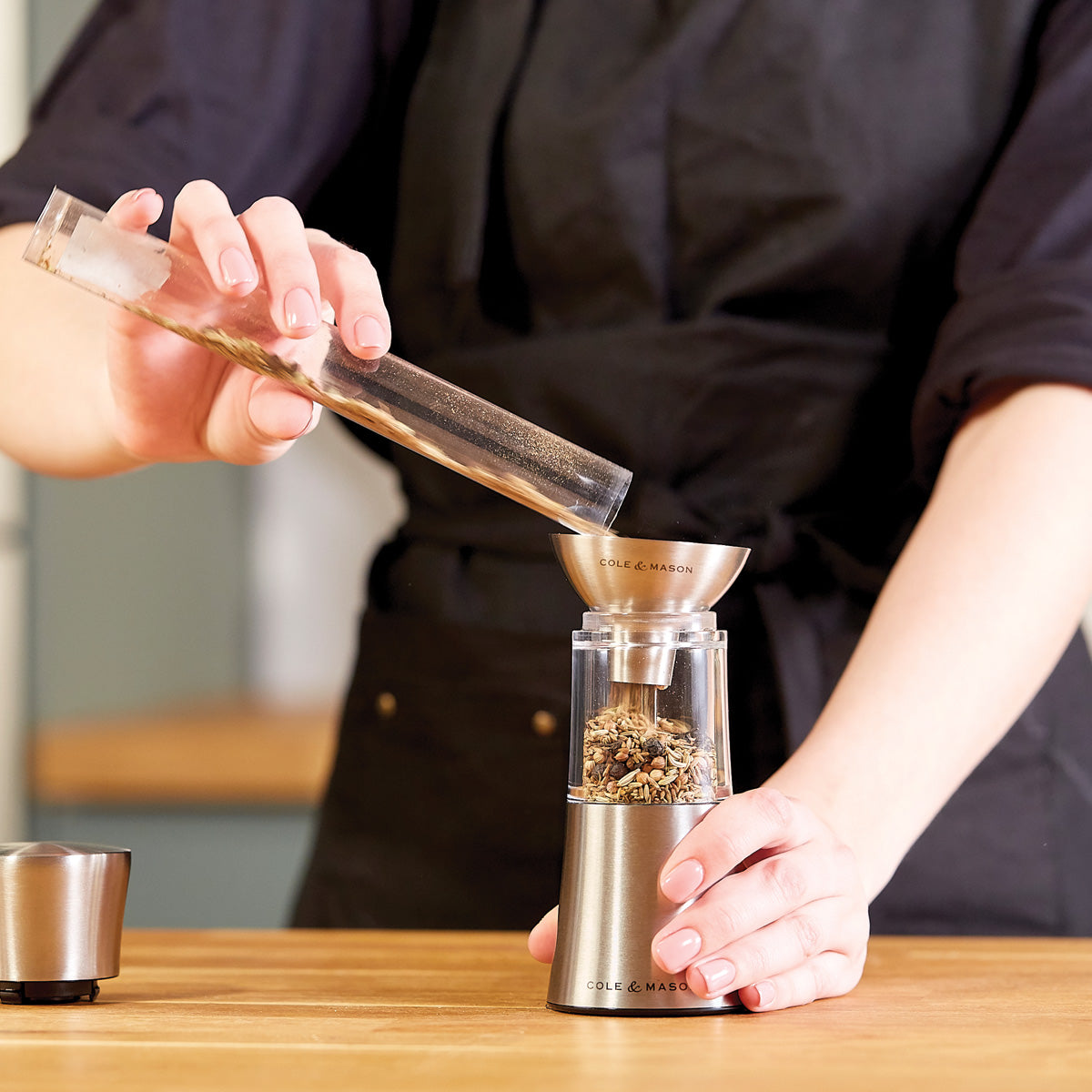 A person in a black apron pours dried herbs from a glass tube into a stainless steel herb grinder on a wooden countertop.