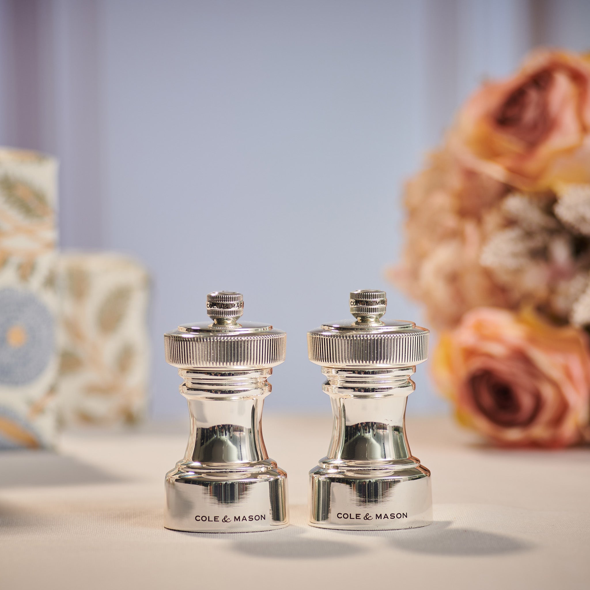 Two shiny silver Cole & Mason salt and pepper mills are displayed side by side on a light surface, with blurred flowers and patterned objects in the background.