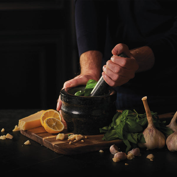#140mm

A person uses a mortar and pestle to grind fresh basil. Nearby, garlic bulbs, lemon slices, Parmesan cheese, and chopped garlic rest on a wooden cutting board. The scene is dimly lit, emphasizing the ingredients.