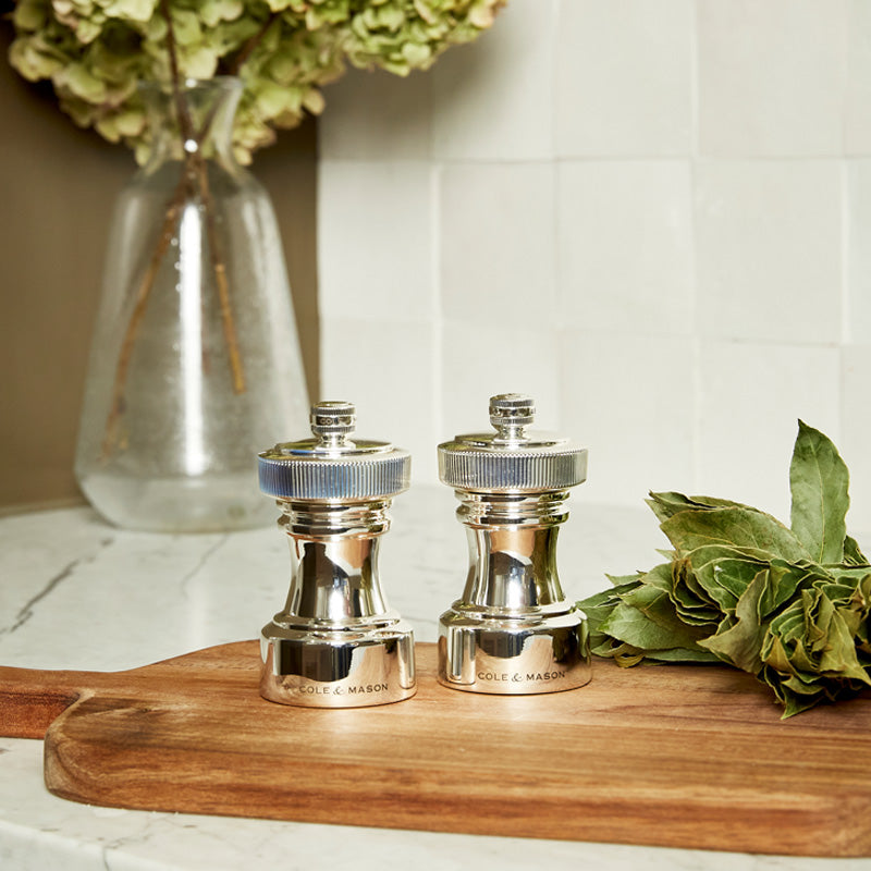 Two silver salt and pepper grinders sit on a wooden cutting board beside bay leaves, with a glass vase of green hydrangeas in the background on a marble counter.