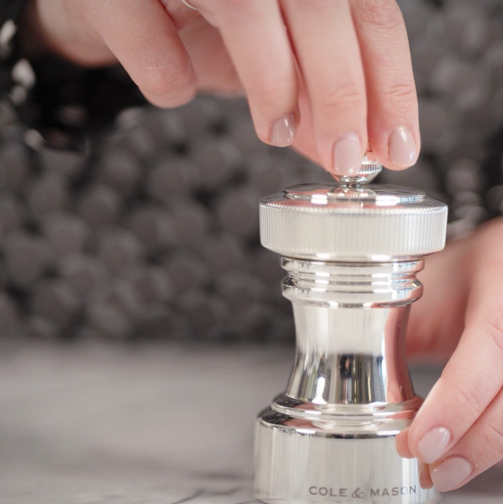 Close-up of a person’s hands using a shiny silver pepper grinder on a light-colored surface, with the brand name Cole & Mason visible at the base. The person has neatly manicured nails.