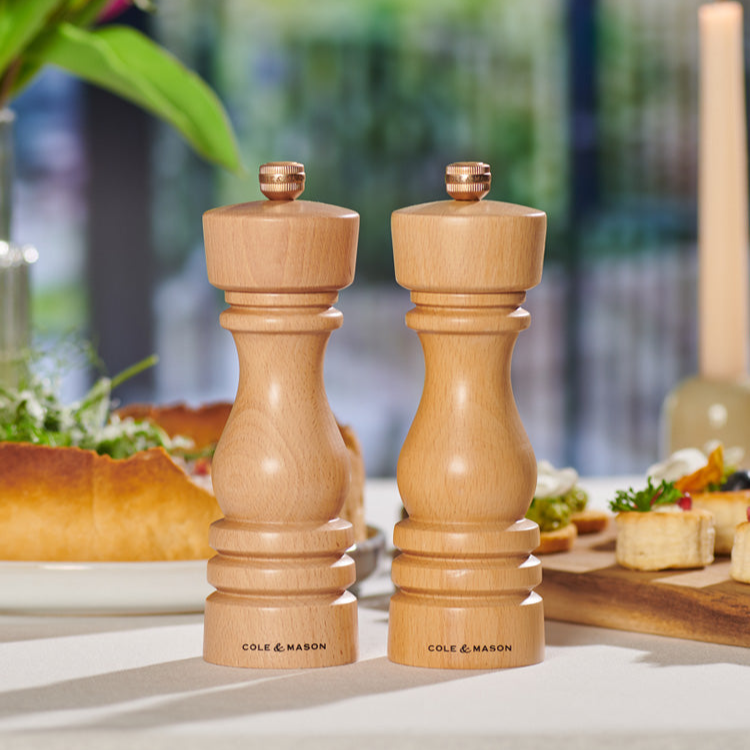 Two wooden Cole & Mason salt and pepper mills are placed side by side on a dining table, with food and greenery blurred in the background.
