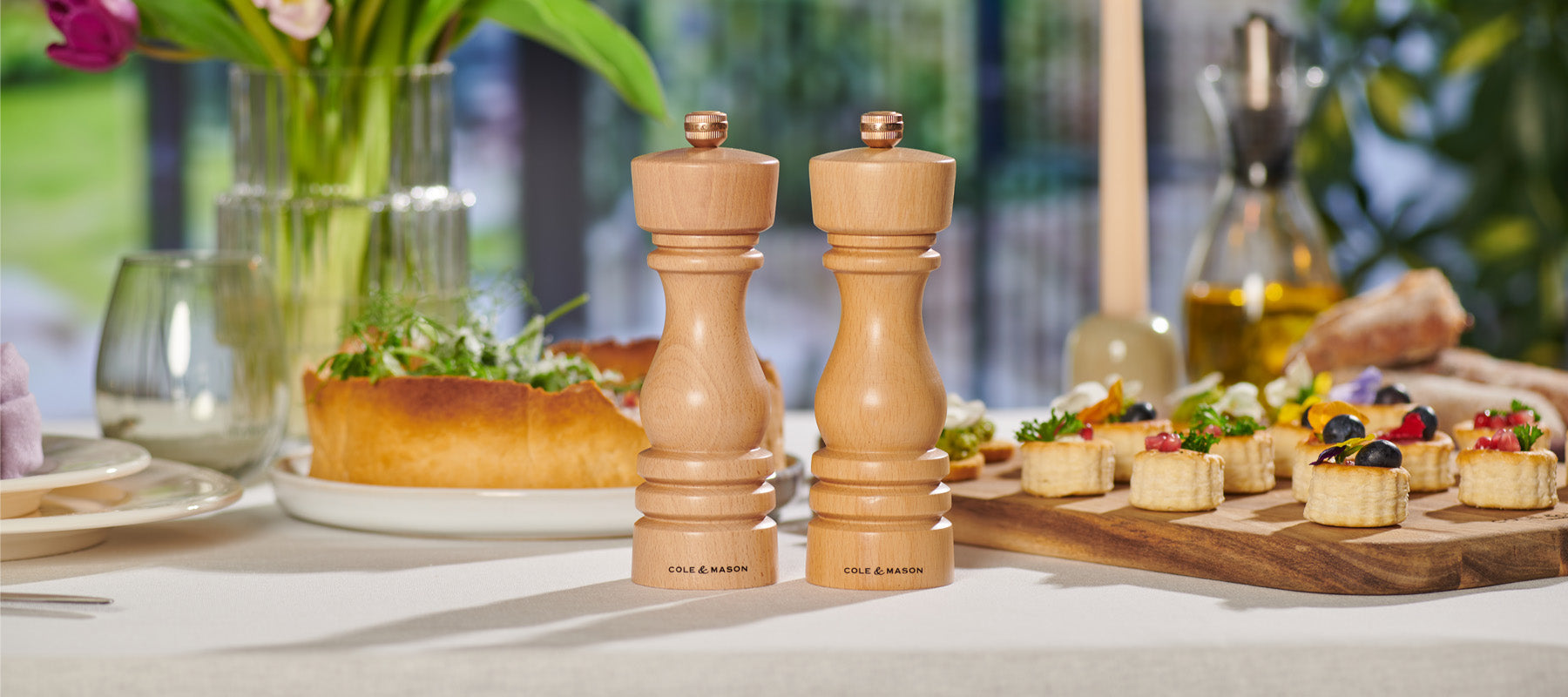 Two wooden salt and pepper mills sit on a set dining table with a salad, appetizers, bread, and flowers in the background. The table is set for a meal in a bright room with natural light.